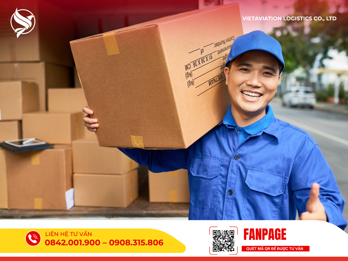 Một nhân viên giao hàng mặc đồng phục xanh đang mỉm cười, vác một thùng hàng carton lớn trên vai.