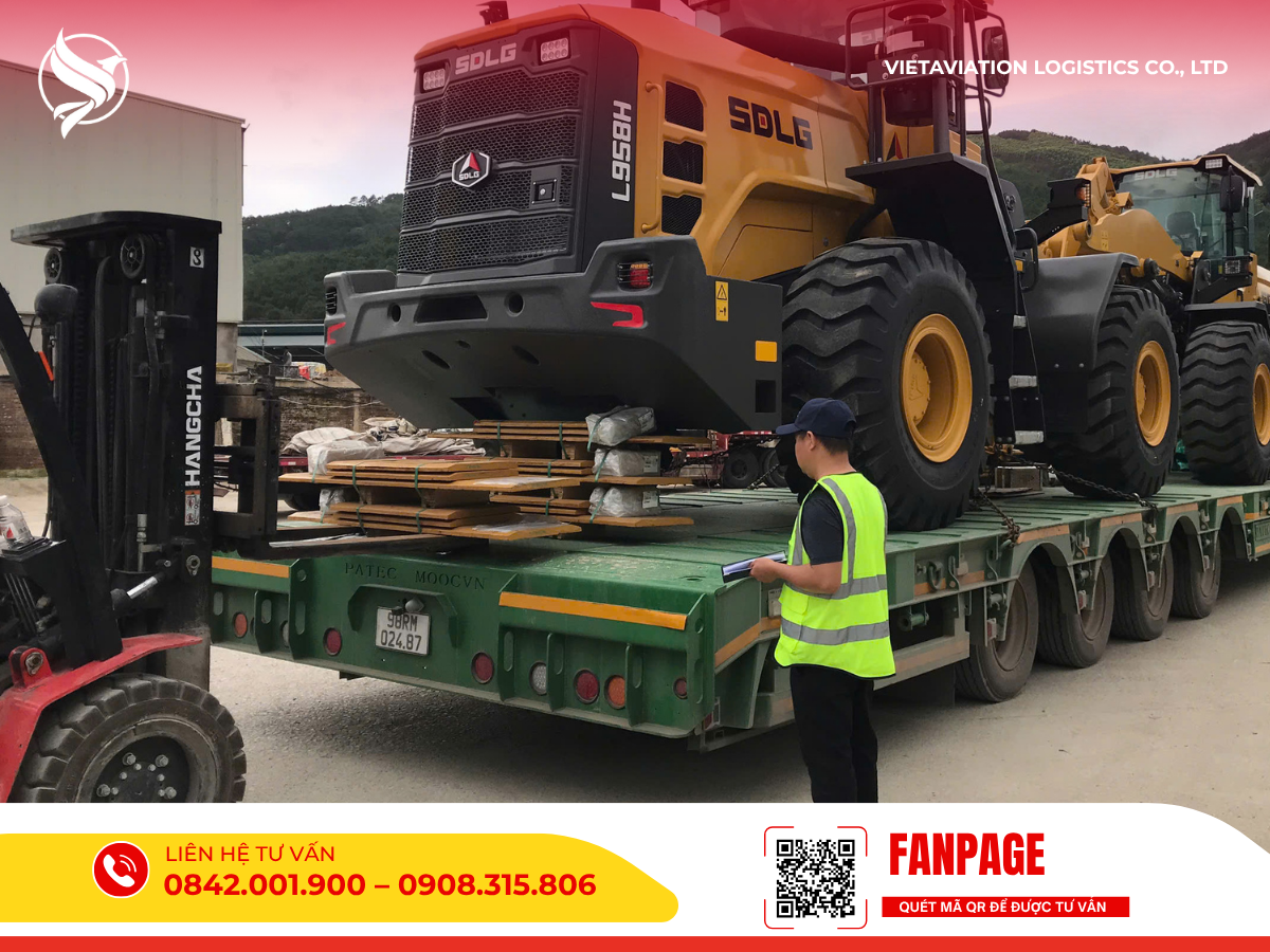 Một nhân viên logistics đang giám sát việc đưa máy xúc lật lớn màu vàng lên xe tải chuyên dụng tại bãi hàng của Vietaviation Cargo.