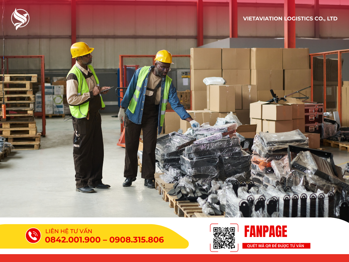Hai nhân viên mặc áo phản quang và đội mũ bảo hộ đang kiểm tra hàng hóa đóng gói trên pallet gỗ bên trong không gian nhà kho rộng lớn.