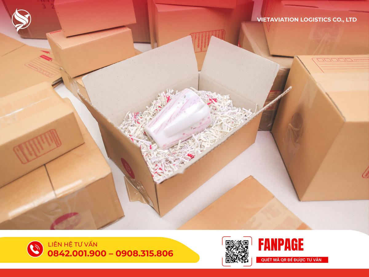 Hình ảnh một chiếc ly sứ họa tiết vân hồng được đặt cẩn thận giữa lớp giấy vụn lót chống sốc bên trong một chiếc thùng carton đang mở nắp, xung quanh là nhiều thùng hàng khác đã được đóng gói và dán băng keo kín.