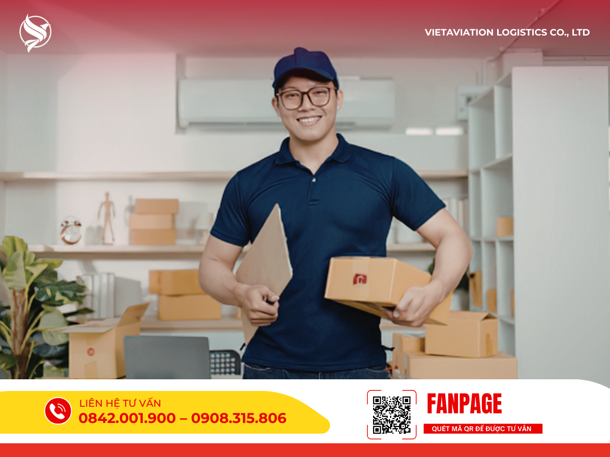 Hình ảnh một nhân viên nam đeo kính, mặc đồng phục áo thun và đội mũ màu xanh đen đang mỉm cười, trên tay cầm một thùng carton nhỏ cùng bìa hồ sơ đứng trong không gian làm việc có nhiều thùng hàng.